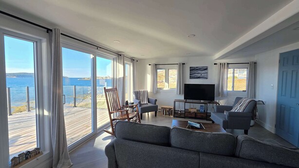 A spacious living room with large windows, a flat-screen TV, and a cozy seating area. - Whales & Trails Vacation Home (Twillingate)
