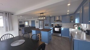 A modern kitchen with blue cabinets, a central island, and a dining area with a table and chairs. - Whales & Trails Vacation Home (Twillingate)