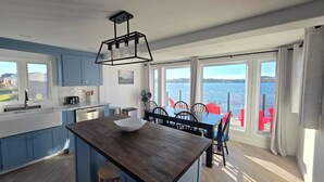 A modern kitchen with a central island, large windows with a view of the sea, and a dining area with red chairs. - Whales & Trails Vacation Home (Twillingate)
