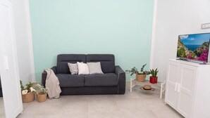 Ein Wohnzimmer mit einem dunkelgrauen Sofa, einem weißen Schrank und einem Fernseher auf einem Lowboard. Übertöpfe und ein Hut stehen auf einem kleinen Tisch.