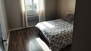 A bedroom with a bed, a window with curtains, a radiator, and a view of buildings outside.