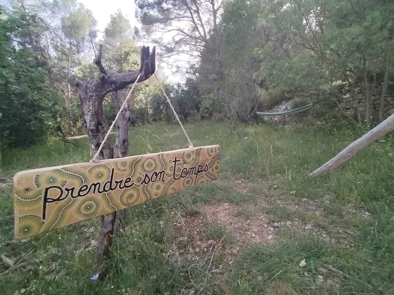 Un letrero de madera con un diseño decorativo y el texto 'Prendre son temps' colgado de un árbol en una zona verde.
