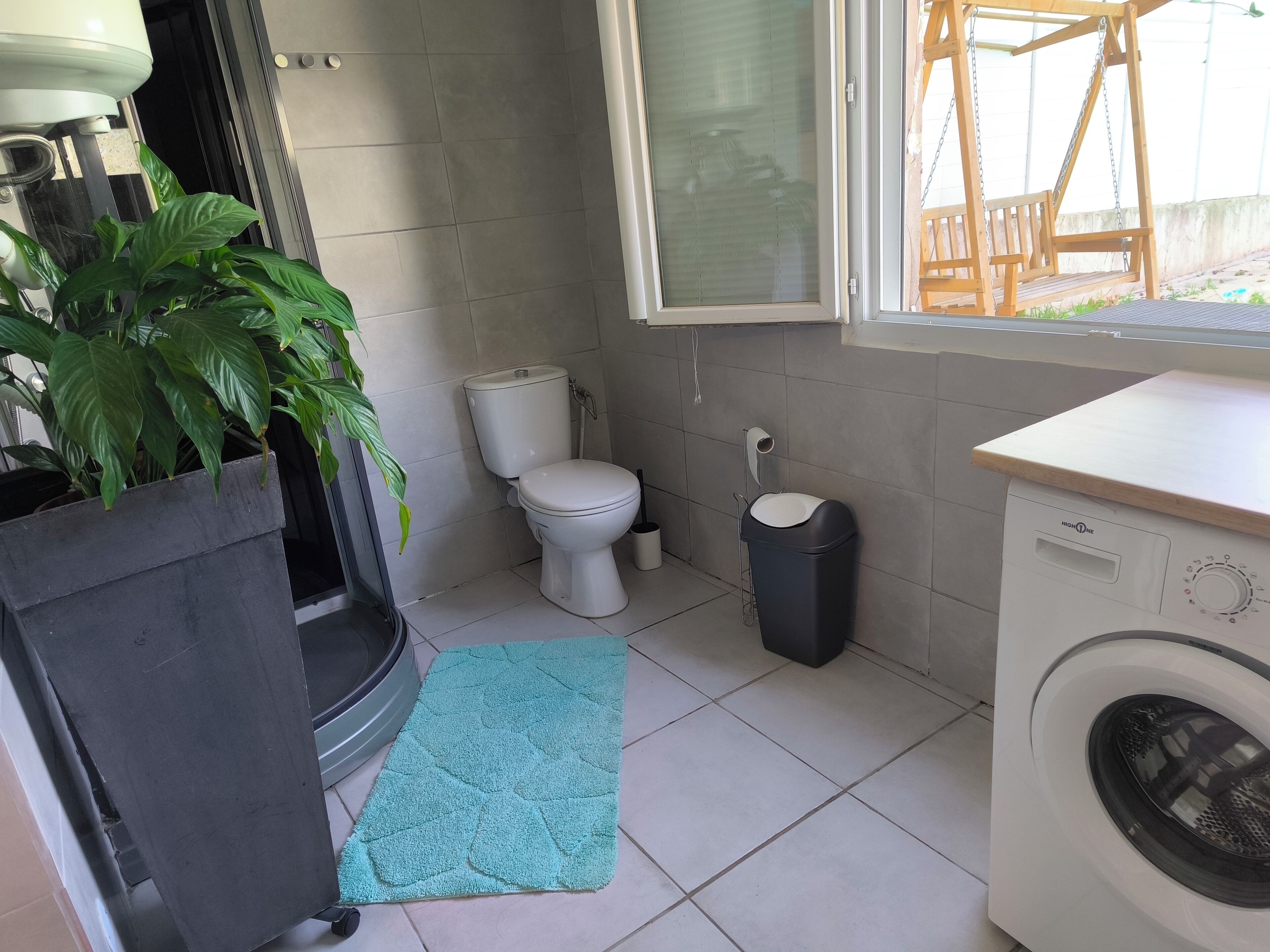 Een compacte, schone badkamer met toilet, prullenbak, wasmachine en een potplante.
