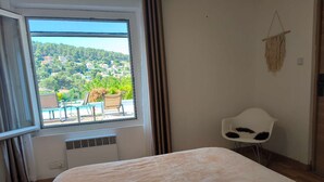 A bedroom with a large bed, a window with a view of a residential area, a chair, and a wall-mounted macrame decoration. - Provence getaway just minutes from Cassis with private pool (Carnoux-en-Provence)
