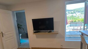 A room with a TV mounted on the wall, wooden flooring, a door leading to a bathroom, and a view of a pool and greenery outside. - Provence getaway just minutes from Cassis with private pool (Carnoux-en-Provence)