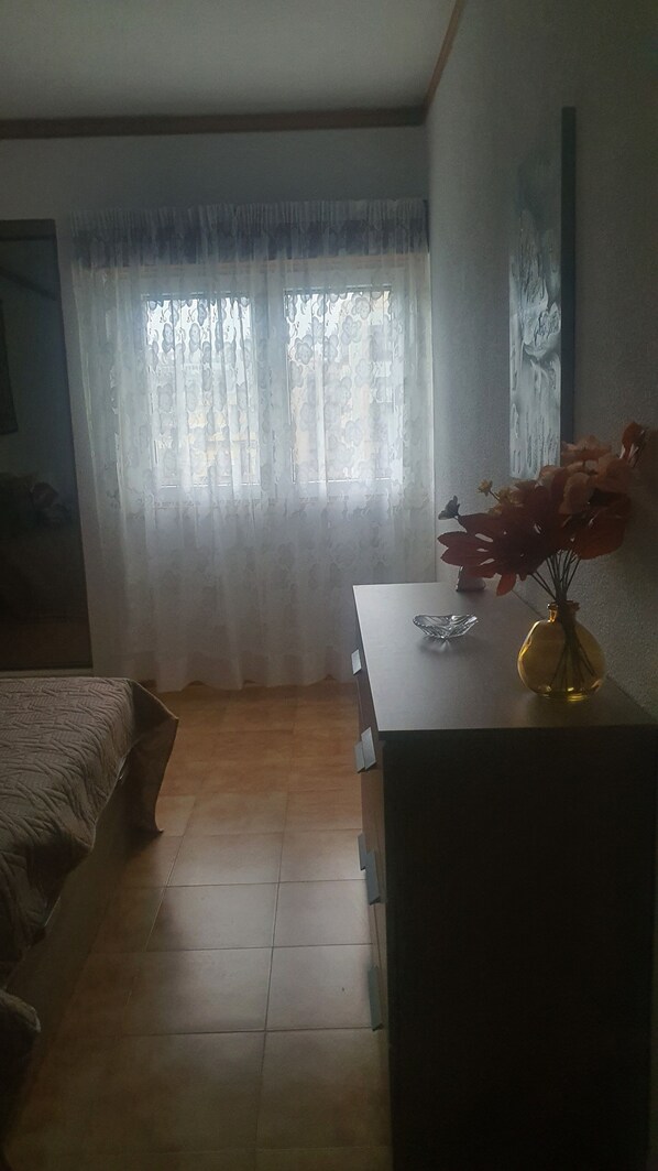 Un dormitorio con una cama, un aparador, un florero con flores y una ventana con cortinas.