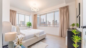 Ein modernes Schlafzimmer mit einem großen Bett, einem Nachttisch mit Lampe, einer kleinen Pflanze und Blick auf die Stadt durch große Fenster.