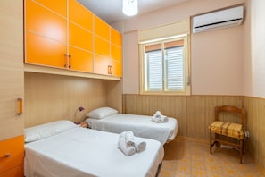 A hotel room with two beds, a wooden wardrobe, a window with blinds, an air conditioner, and a chair with a towel.