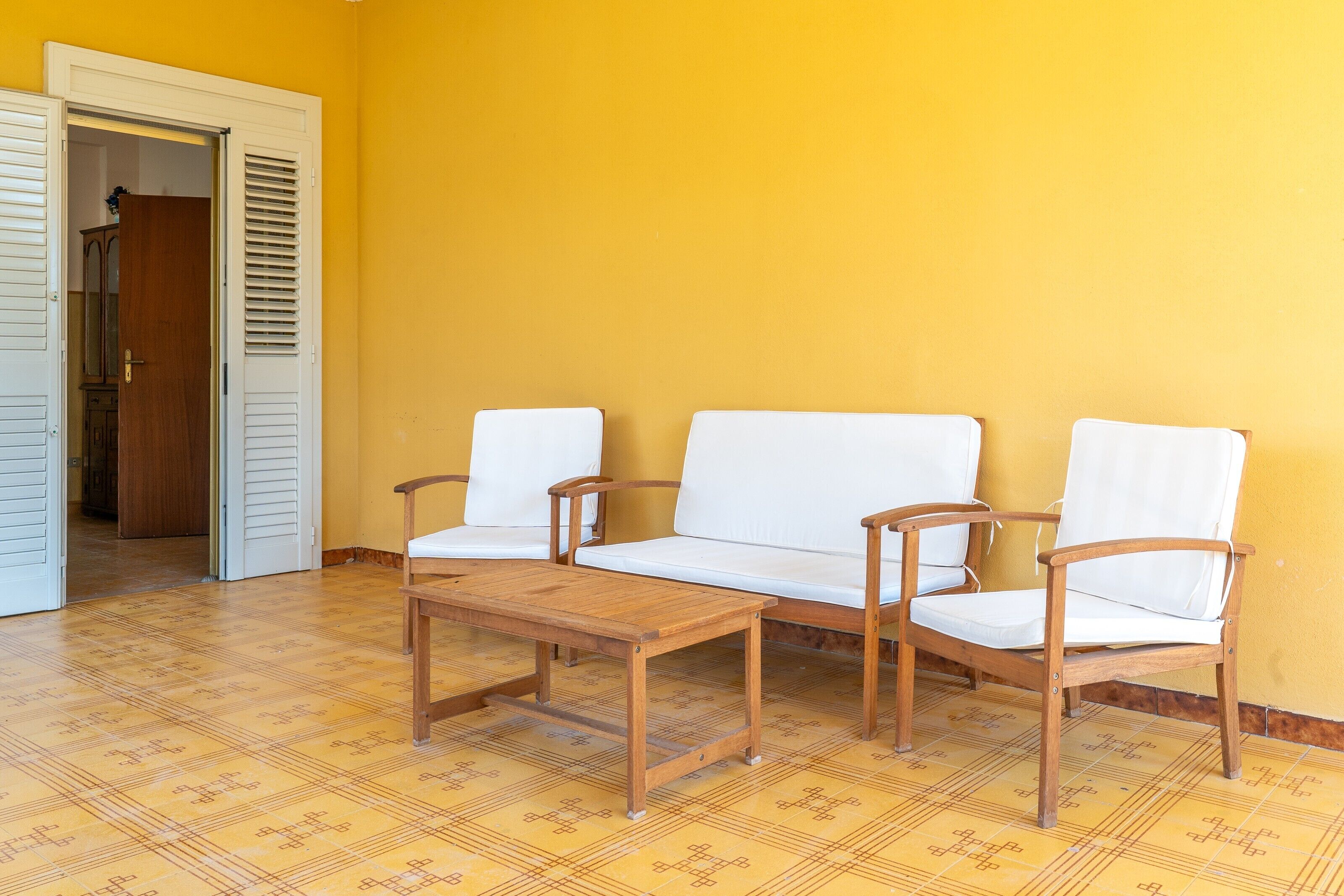 Een kamer met een gele wand, een houten vloer, een witte deur en een zithoek met een houten tafel en twee stoelen.