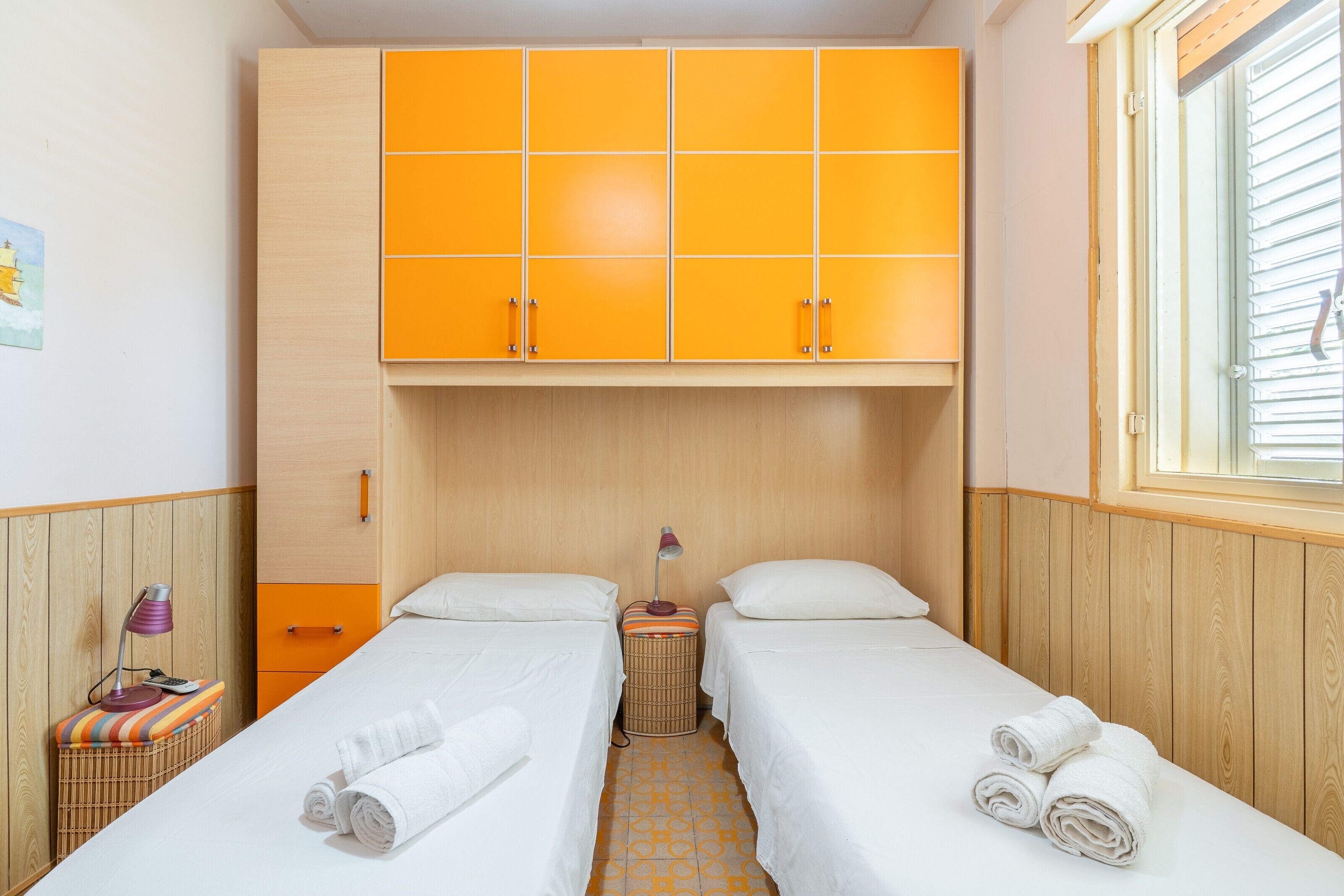 Una habitación con dos camas, un armario de madera, una ventana y una mesita de noche con lámpara.