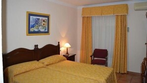 Habitació d'hotel amb llit doble, una nit i una làmpada, una cadira, una finestra amb cortines i un quadre al mur.