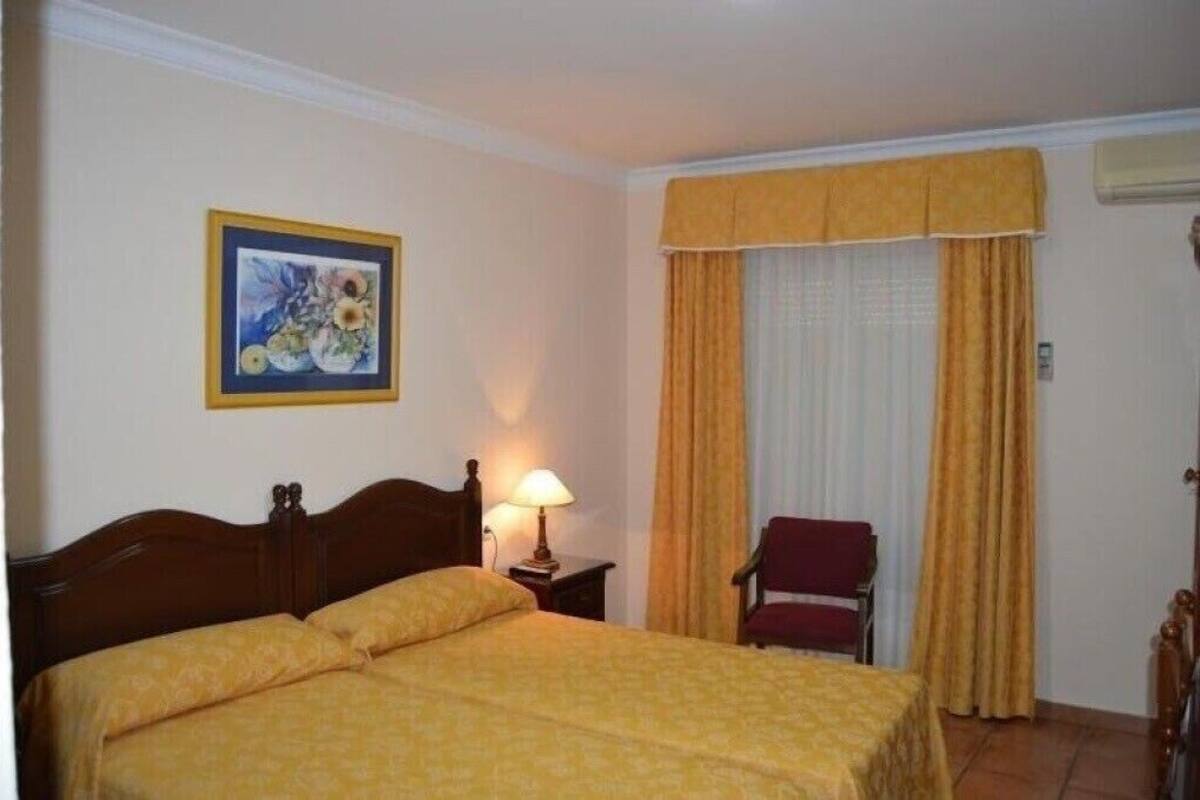 A hotel room with a double bed, a nightstand with a lamp, a chair, a window with curtains, and a framed picture on the wall.