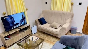 A living room with a TV, a sofa, a glass coffee table, and a grey armchair. - The Harmony Oasis Hotel and Suites (Port Harcourt)