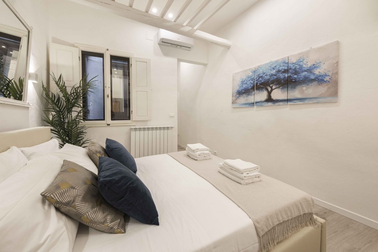 Een slaapkamer met een bed, kussens, een radiator, een raam met luiken en een schilderij aan de muur.