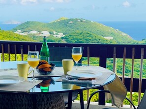 A table with a glass of orange juice, a glass of white wine, a bottle of water, and a bowl of fruit on a balcony with a view of green hills and the sea.