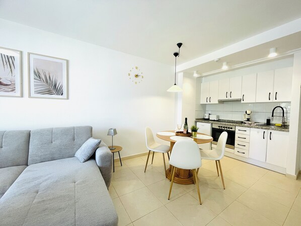 Een moderne woonkamer met een grijze bank, een eettafel met stoelen en een keuken met witte kastjes en een fornuis.