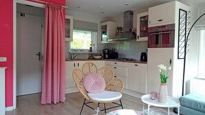 A modern kitchen with white cabinets, a stove, and a sink. There is a wicker chair with a cushion and a small white table with a vase.