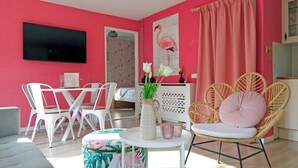 A living room with a TV, white chairs, a wicker chair with a cushion, a small table with a vase and glasses, and a pink wall.