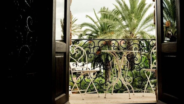 Une porte ouverte donnant sur un balcon orné d’une balustrade en fer forgé, avec des palmiers en arrière-plan.