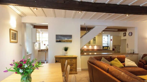 A cozy living room with a brown leather sofa, a wooden dining table with a vase of flowers, and a staircase leading to a kitchen area.