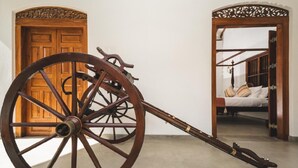 Une grande roue en bois, dotée d’un long manche, probablement un objet historique, se trouve dans une pièce avec une porte en bois et un lit recouvert de draps blancs.