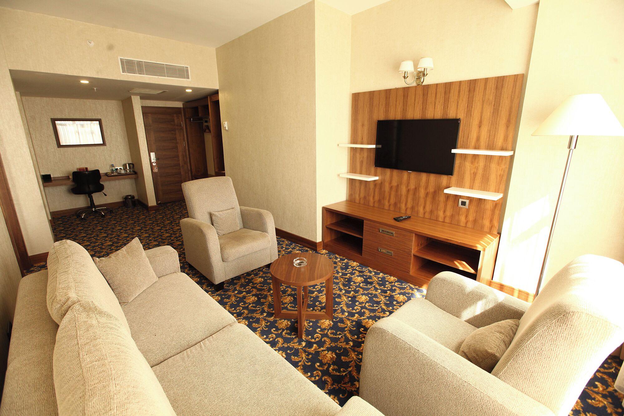 A hotel room with a sofa set, a TV mounted on a wooden wall, a small table, and a lamp.