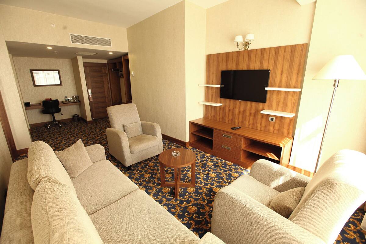 A hotel room with a sofa set, a TV mounted on a wooden wall, a small table, and a lamp.