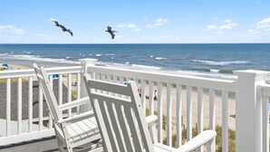 A beach view from a deck with white railings and chairs, overlooking the ocean and beachgoers.