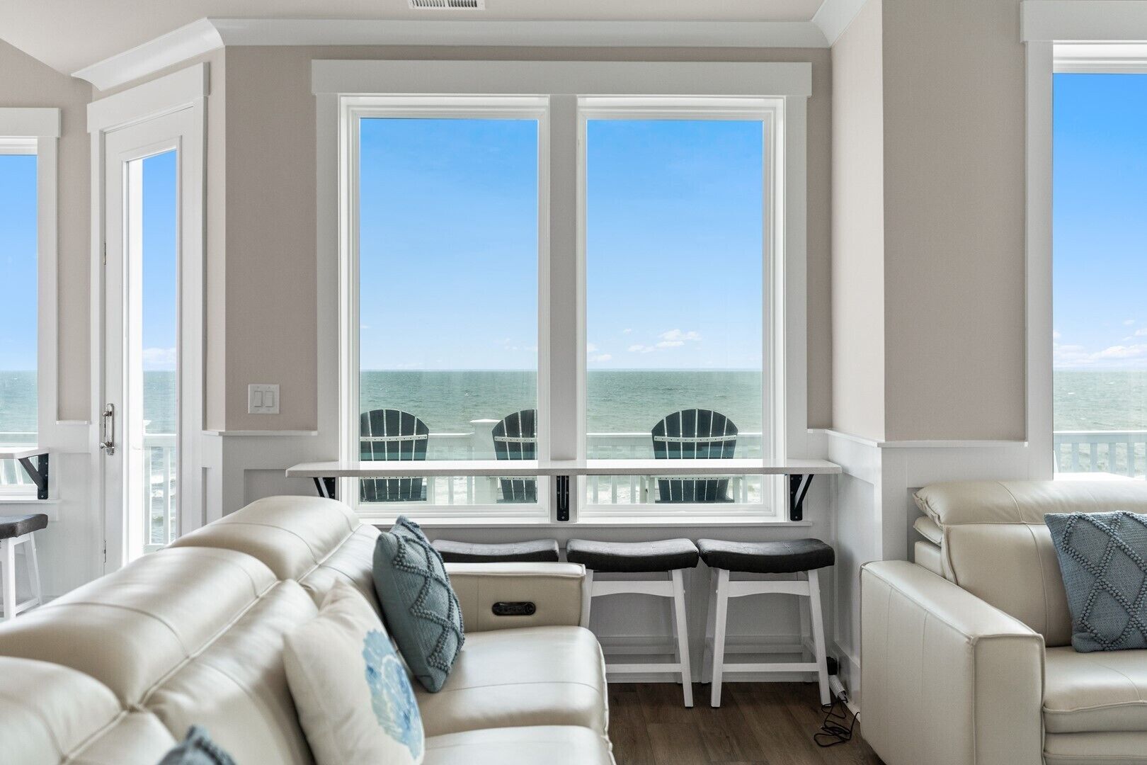 Een kamer met een groot raam met uitzicht op de oceaan, een witte bank, een klein tafeltje en twee stoelen.