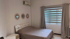 Un dormitorio con cama, mesita de noche, lámpara, ventana con cortinas y aire acondicionado instalado en la pared.