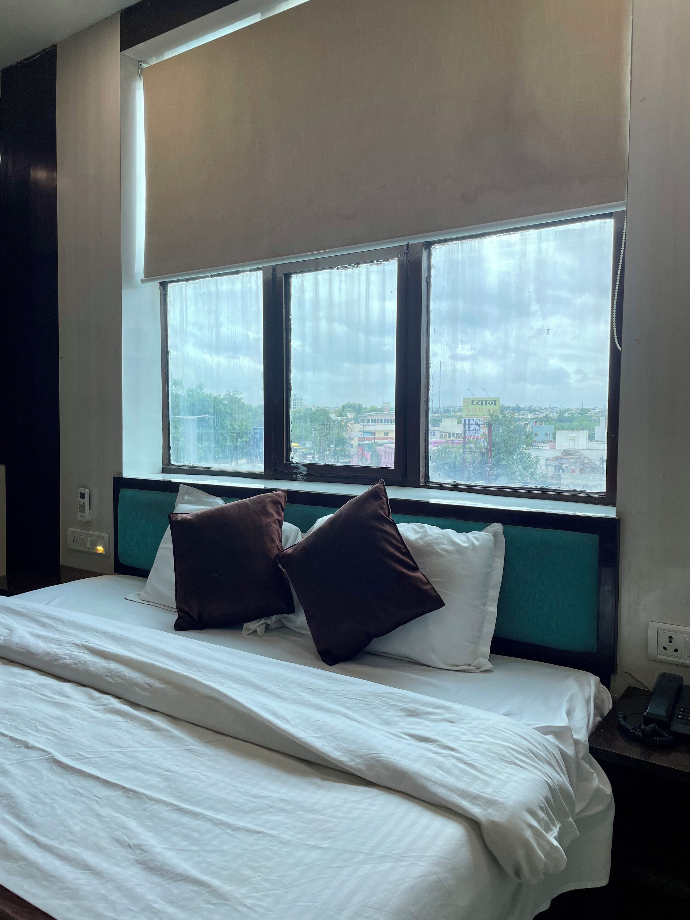 Habitación de hotel con cama, ventana con persianas y vista a la ciudad.