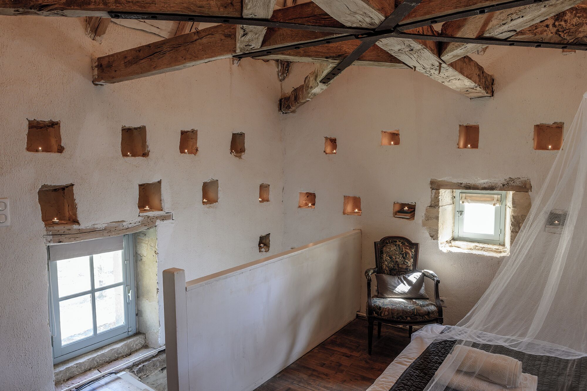 A room with exposed wooden beams, a chair, a bed, and multiple small windows.