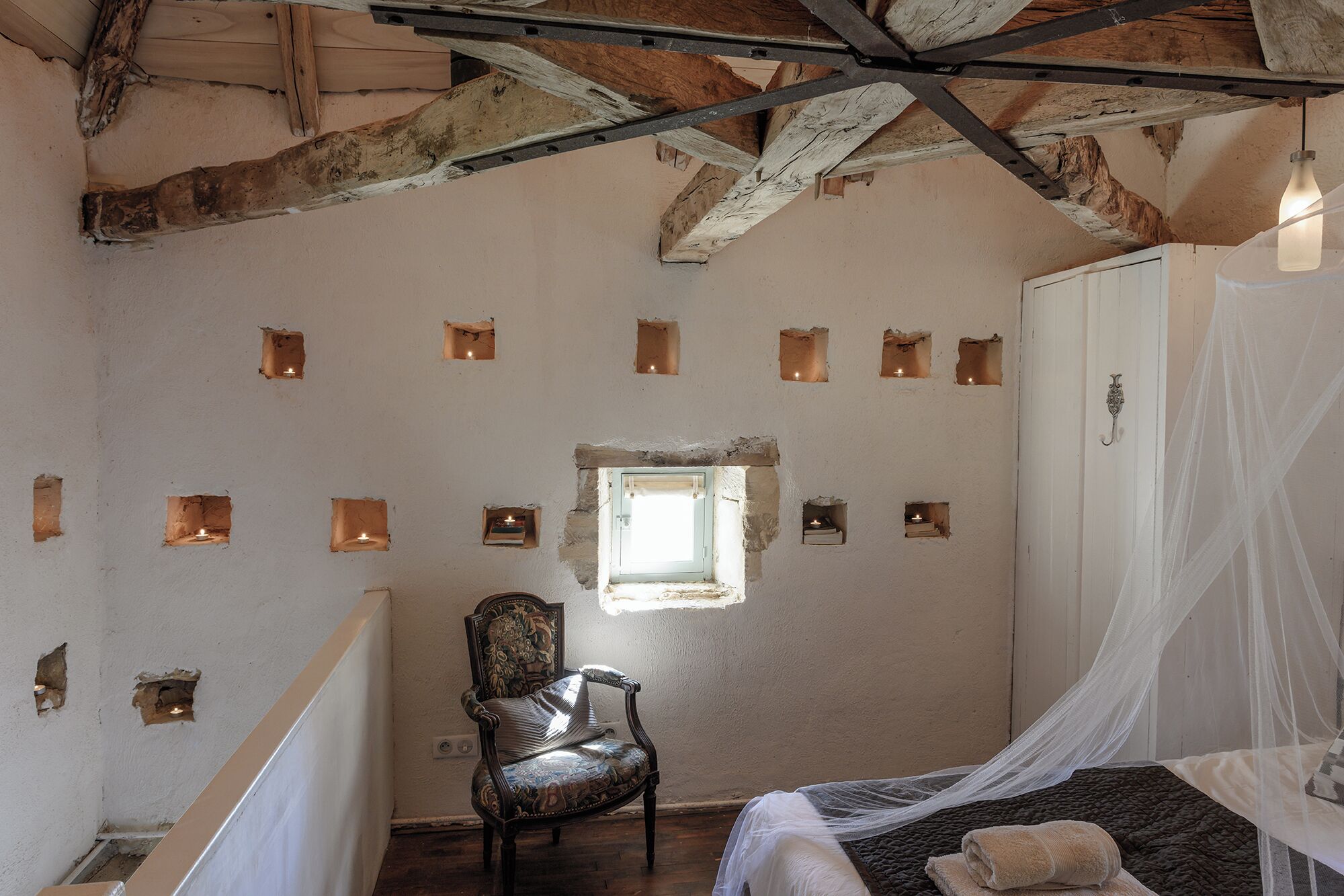 Ein Schlafzimmer mit rustikalem Holzbalken, einem Holzstuhl, einem Bett mit Baldachin und mehreren kleinen Fenstern.