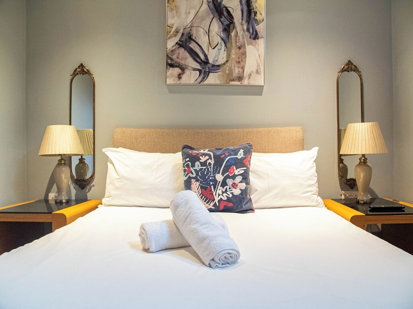 Una cama bien hecha con edredón blanco, dos almohadas y una toalla enrollada. Hay dos mesitas de noche con lámparas y un cuadro abstracto grande en la pared.