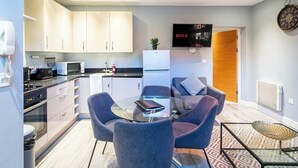 A modern kitchen with white cabinets, a microwave, and a coffee machine. There is a dining area with a glass table and two chairs. A television is mounted on the wall, and a clock is visible on the wall. - Spacious Ensuite 2-Bedroom Flat in Kelham Island (South Yorkshire)