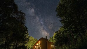 A house with a large window and a chimney, surrounded by trees under a starry night sky. - Designer Forest Cabin w/Hot Tub Near CherrySprings (Coudersport)