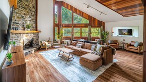 A spacious living room with a stone fireplace, large windows, and wooden flooring. - Designer Forest Cabin w/Hot Tub Near CherrySprings (Coudersport)