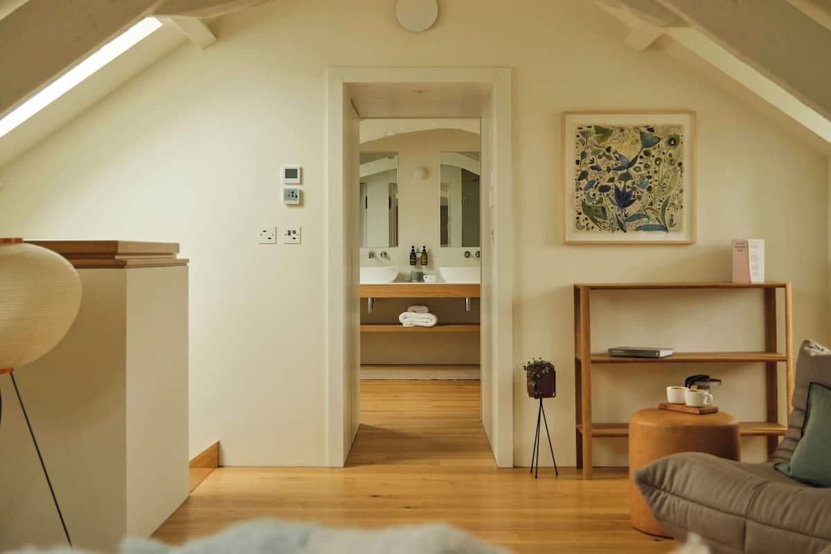 Ein gemütliches Schlafzimmer im Dachgeschoss mit schrägen Wänden, Holzboden, einem Bett, einem Schreibtisch, einem Stuhl, einem kleinen Tisch mit Pflanze und einem Bild an der Wand.