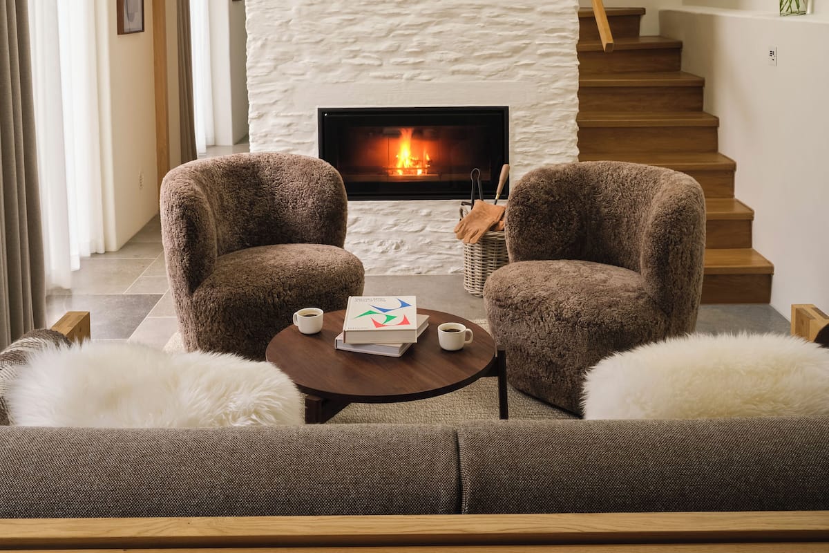 Ein modernes Wohnzimmer mit Kamin, zwei Sesseln, einem Couchtisch mit Büchern und Tassen sowie einer Holztreppe.
