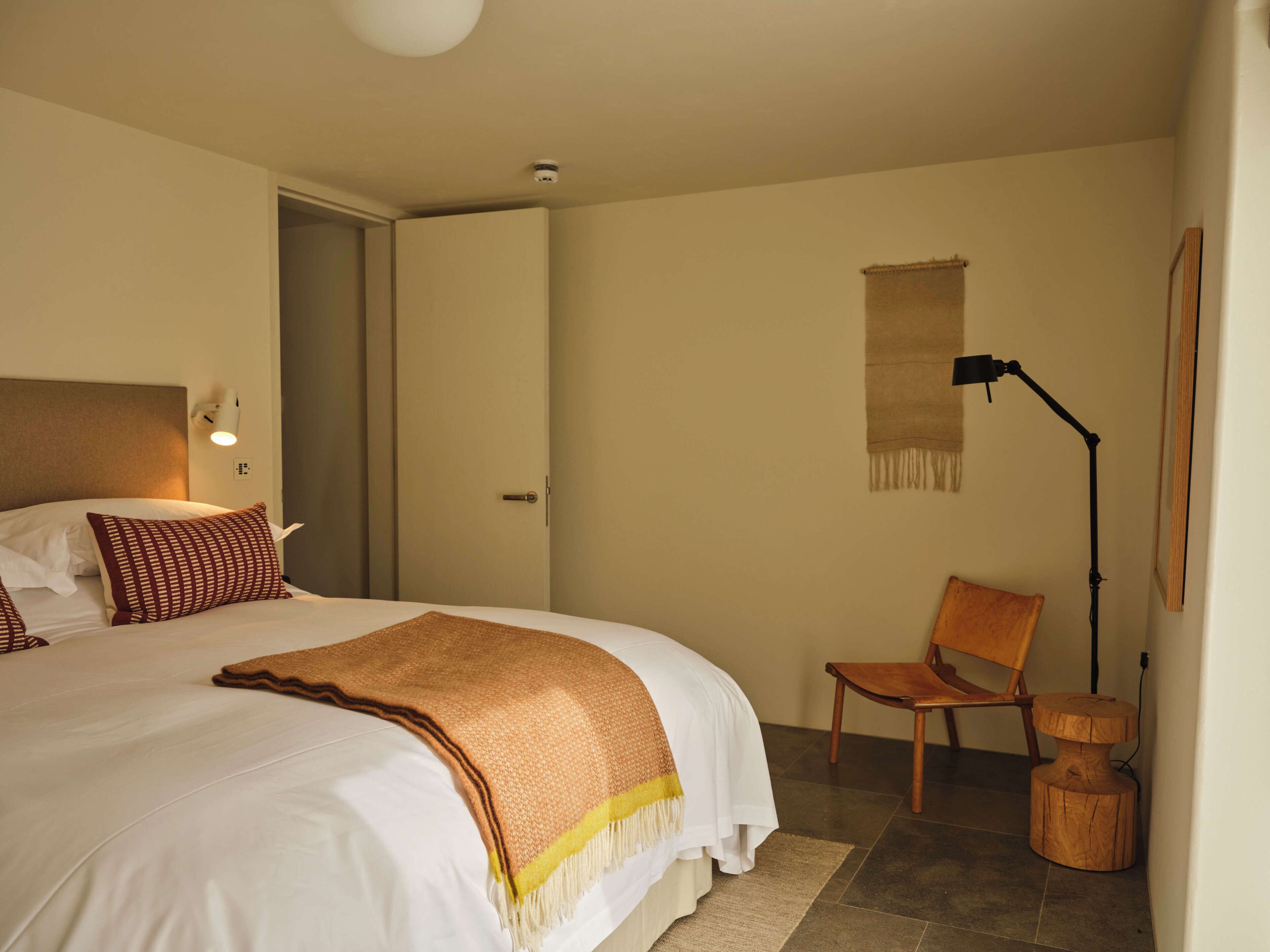 Hotellihuone, jossa on sänky, tuoli, lamppu ja pieni puujakkara.