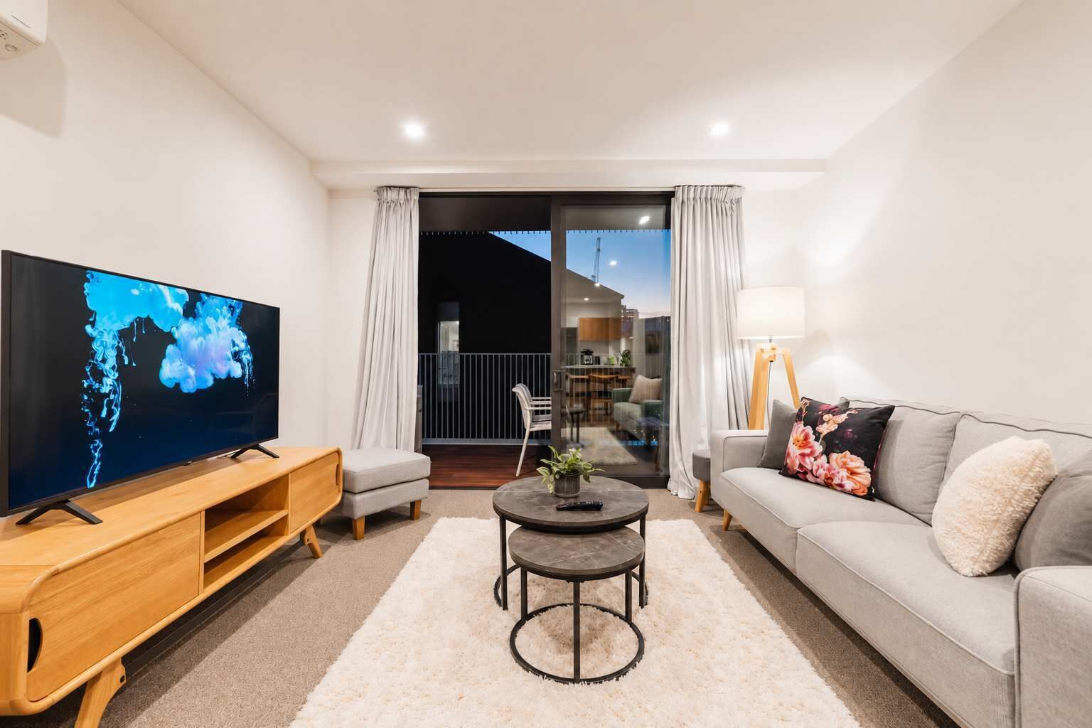A modern living room with a flat-screen TV, a grey sofa, a wooden TV stand, a round coffee table, and a balcony with a railing.