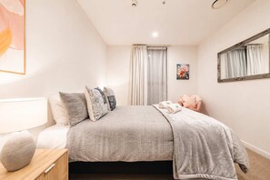 A bedroom with a bed, pillows, a bedside table, a lamp, a mirror, and a window with curtains.