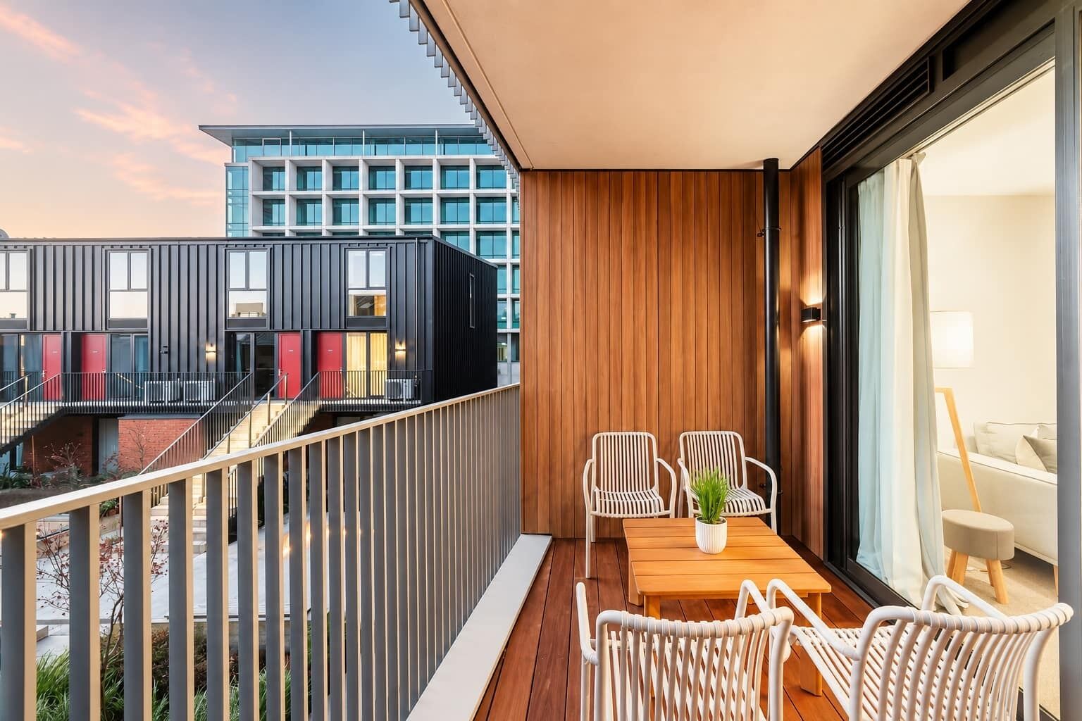 A balcony with a wooden table and white wicker chairs, overlooking a modern building with large windows.