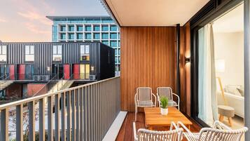 A balcony with a wooden table and white wicker chairs, overlooking a modern building with large windows.