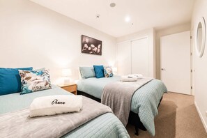 A hotel room with two beds, each with a blue bedspread and matching pillows, a bedside table with a lamp, and a framed picture on the wall.