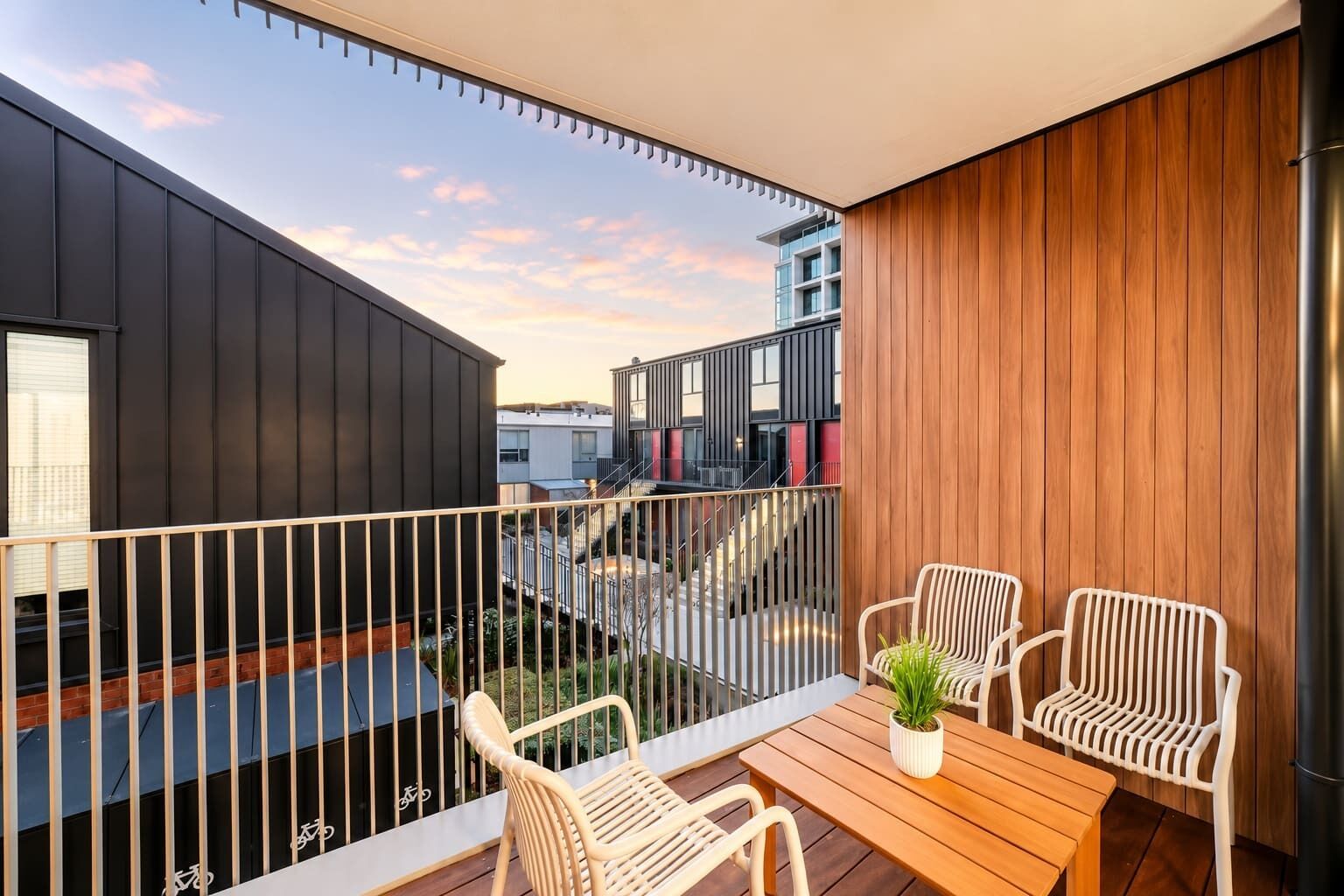 A balcony with a wooden table and chairs, a potted plant, and a view of adjacent buildings.
