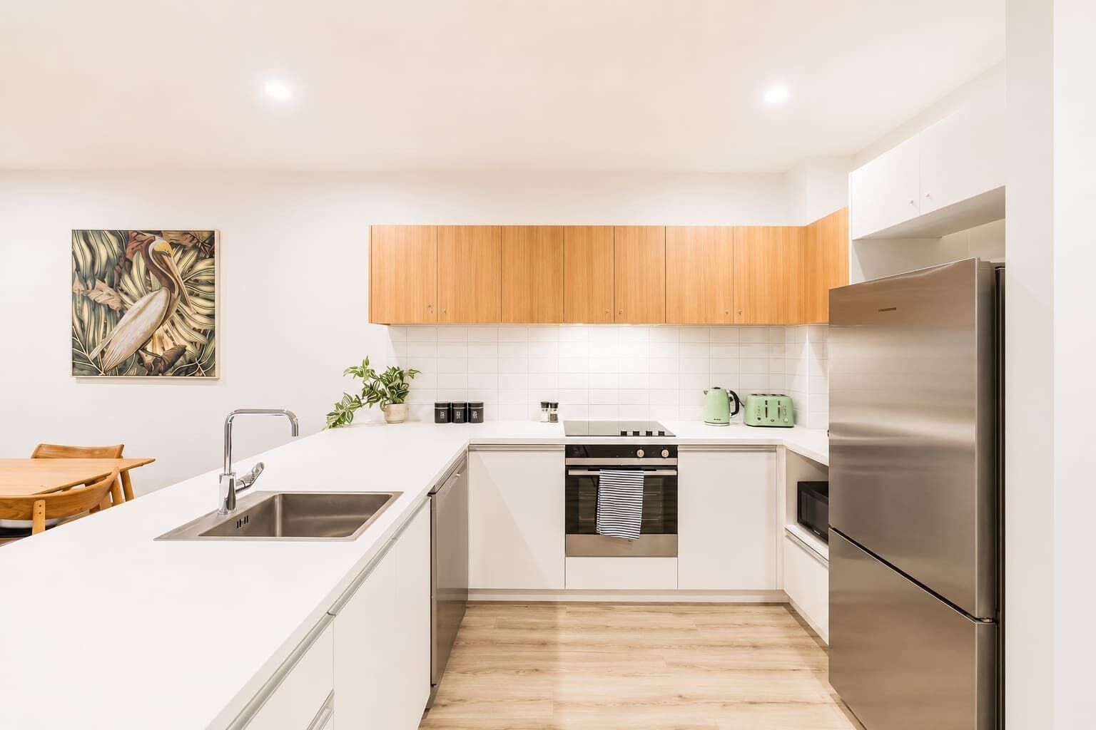 A modern kitchen with white cabinetry, a stainless steel refrigerator, a built-in oven, and a dining area with a wooden table.