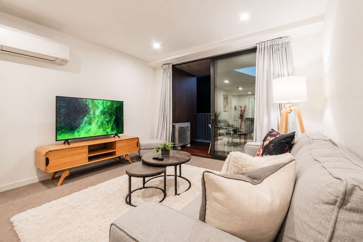 A modern living room with a flat-screen TV, a wooden entertainment unit, a round coffee table, a sofa, and a glass door leading to a balcony.