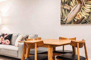 A dining area with a wooden table and chairs, a grey sofa with floral cushions, and a wall painting.