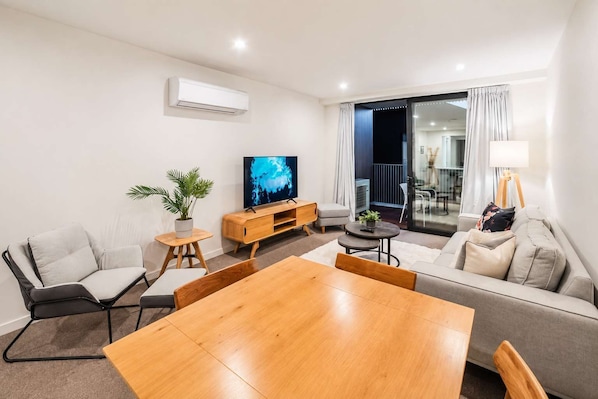 A modern living room with a wooden dining table, sofas, a TV, and a balcony with curtains.
